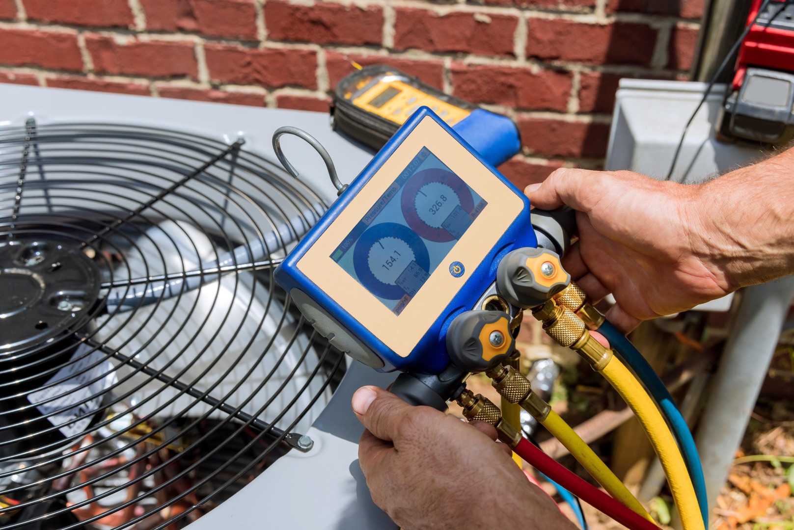 A person uses a digital gauge manifold to check refrigerant levels on an outdoor air conditioning unit next to a brick wall, perfect for a landing page header showcasing HVAC expertise.