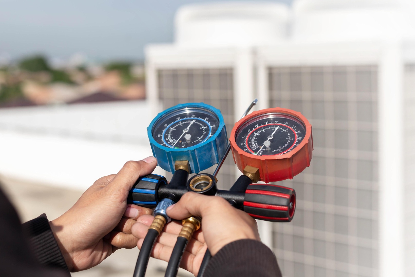 Person holding a manifold gauge set with blue and red dials, measuring pressure in HVAC systems—perfect for your landing page header, with outdoor equipment visible in the background.