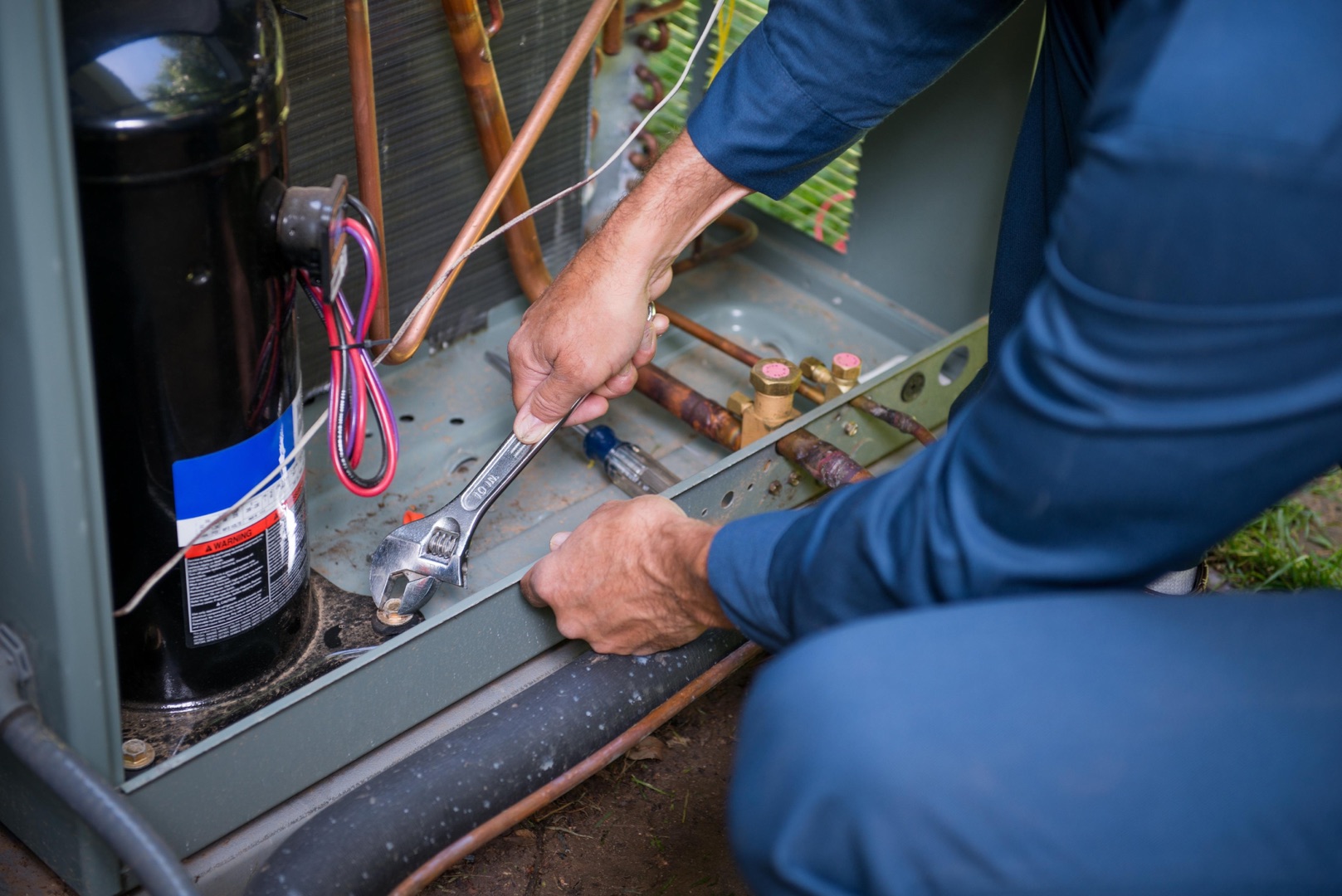 A person uses a wrench to tighten a bolt on the exterior unit of an air conditioning system, perfect for a landing page or header showcasing HVAC maintenance services.