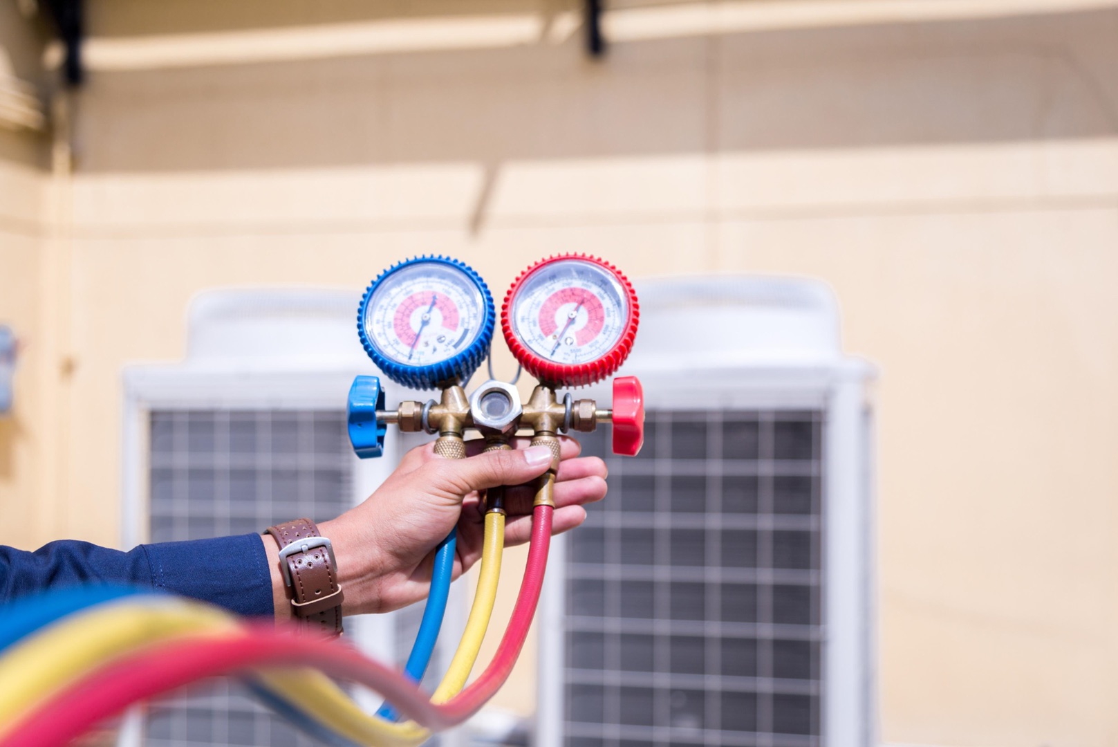A person holds HVAC manifold gauges with red, blue, and yellow hoses, in front of an outdoor air conditioning unit—perfect for a landing page header image.