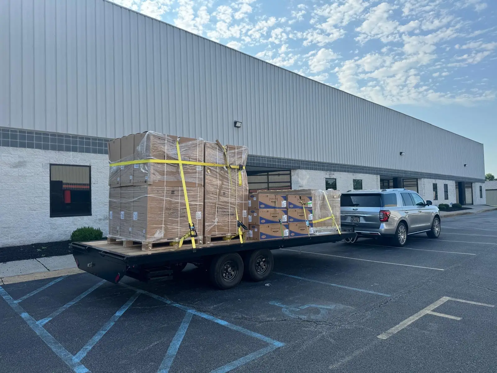 A silver SUV, possibly belonging to an air conditioning repairman, tows a flatbed trailer loaded with large, shrink-wrapped pallets and boxes outside a warehouse building.